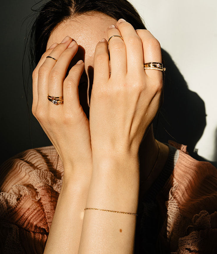 Potrait einer jungen Frau im Licht, die zärtlich ihr Gesicht mir ihren Händen verdeckt, die mit vielen nachhaltigen Goldringen geschmückt sind