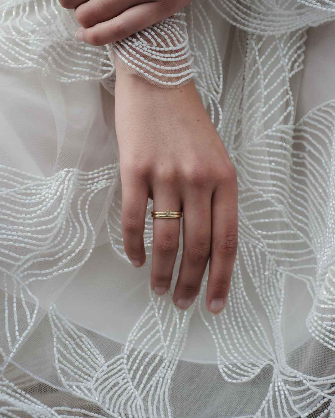Ein Model hält ihre and mit Eheringen aus recyceltem Gold geschmückt von oben ins Bild vor einem weißen Kleid als Hintergrund