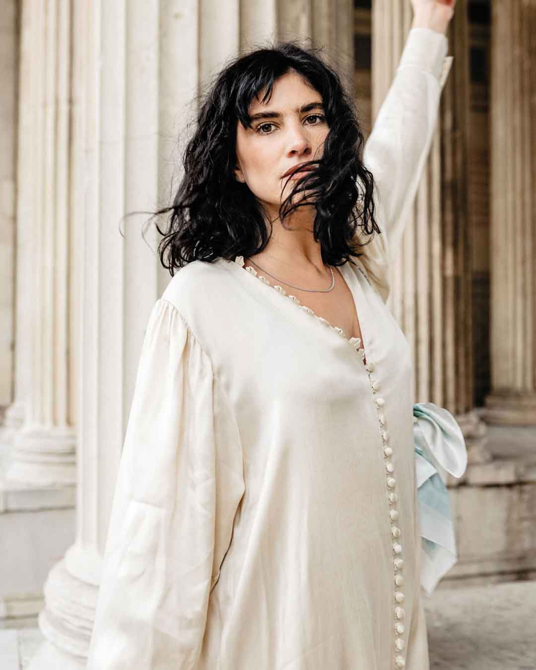 schwarzhaariges Model in schlichtem weißen Kleid steht in einer Säulenhalle und präsentiert die achtsame Halskette von Maren Jewellery