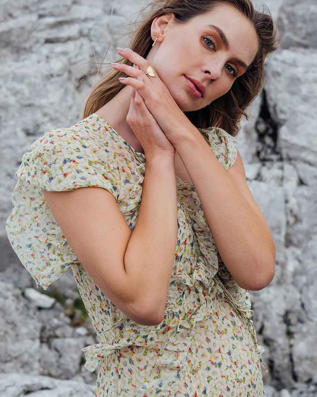 Ein junges Model im zarten Blumenkleid vor felsigem Hintergrund schaut in die Kamera und präsentiert nachhaltige Ringe und Ohrstecker von Maren Jewellery