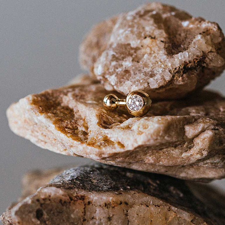 Kugelohrring mit zwei Kugeln aus recyceltem Gold und einem Labordiamant von Maren Jewellery wird auf einem Stapel rauer Steine präsentiert