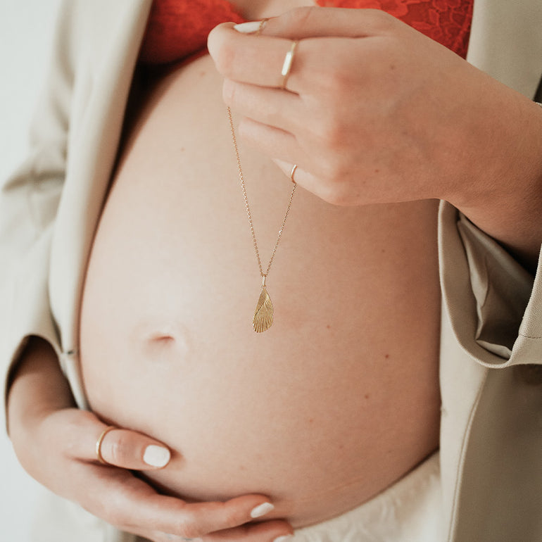 Frau mir Schwangerschaftsbauch hält Goldkette mit Storchenflügel Anhänger zur Geburt