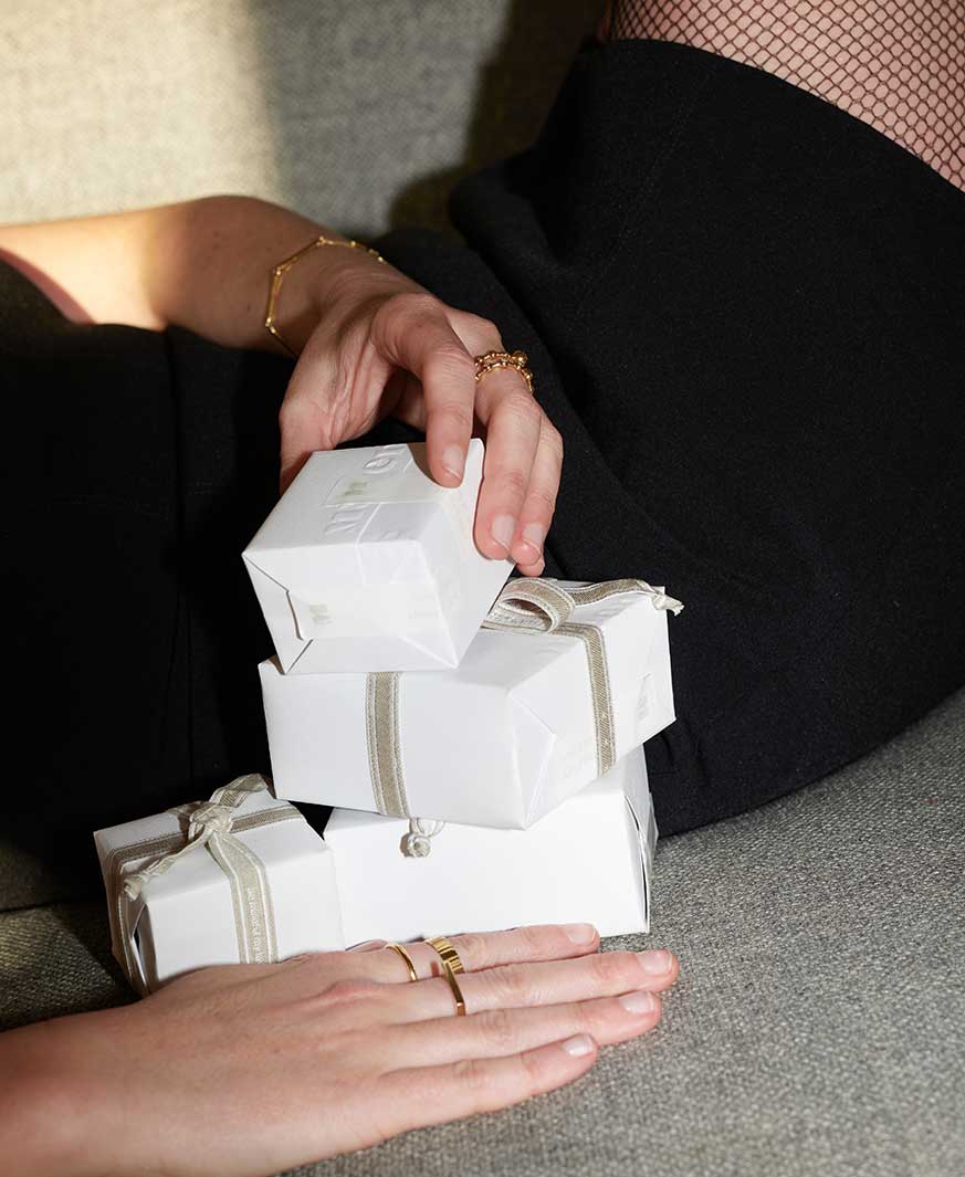 Bildausschnitt einer jungen Frau im schwarzen Kleid und mit nachhaltigem Goldschmuck, die Geschenkverpackungen für Schmuck vor sich stapelt.