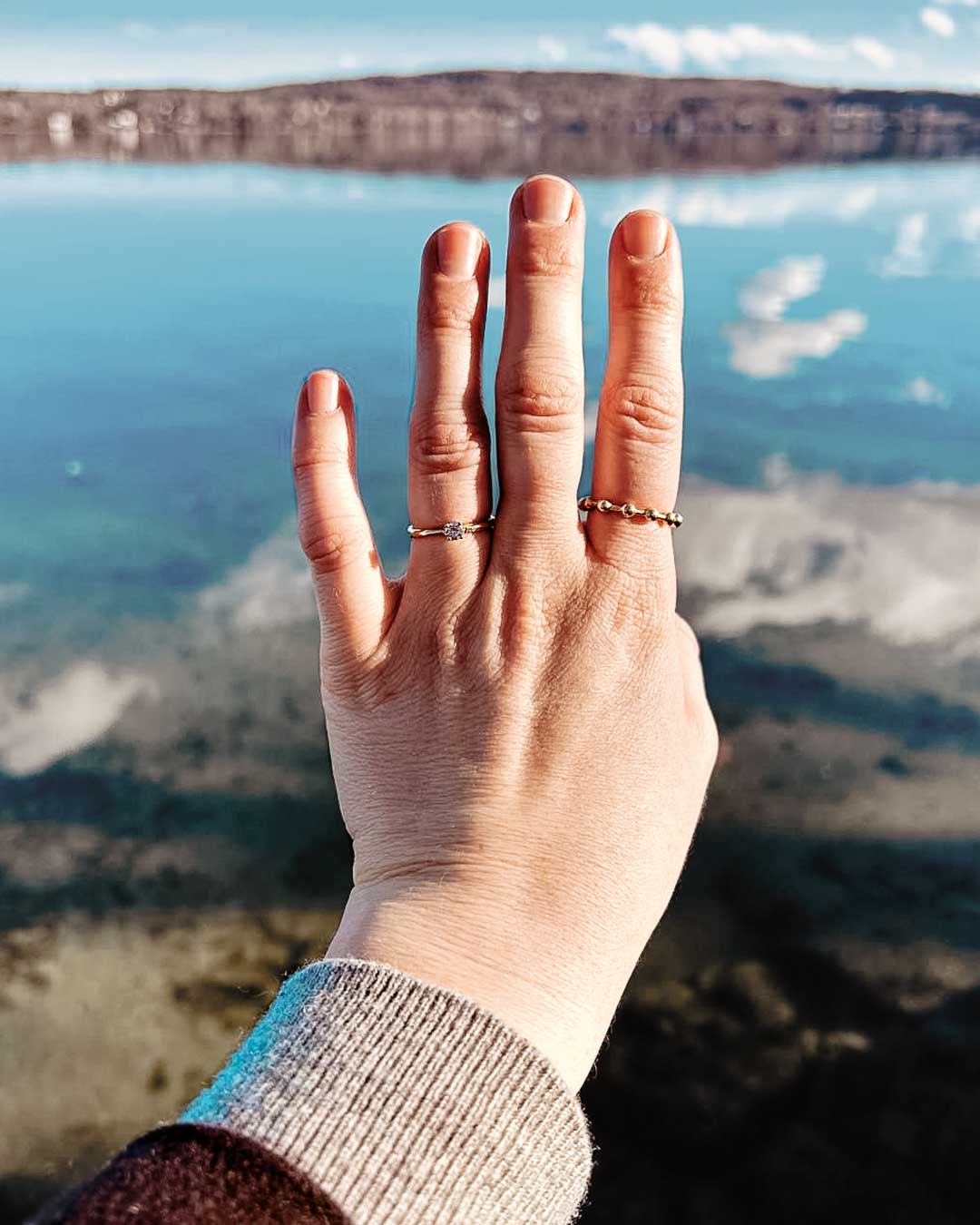 Satisfied customer holds up her hand with custom-made rings by Maren Jewellery in front of a reflective lake toward the camera
