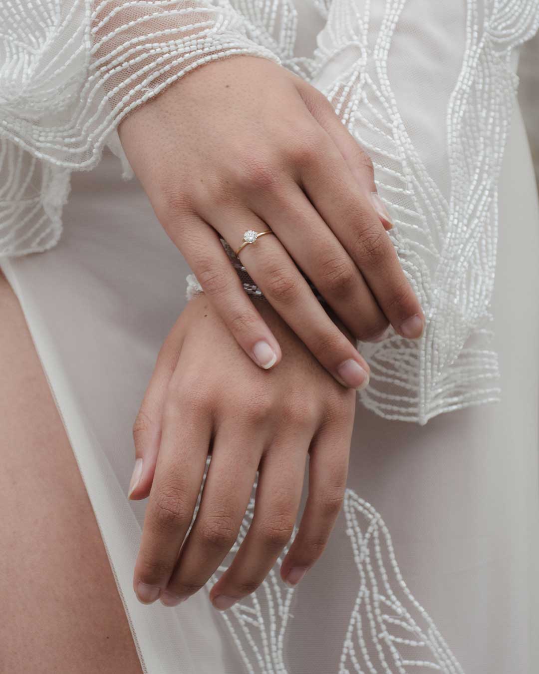 Bild zeigt zarte Frauenhände mit einem nachhaltigen Verlobungsring aus recyceltem Gold und Labordiamant von Maren Jewellery vor einem weißen Kleid