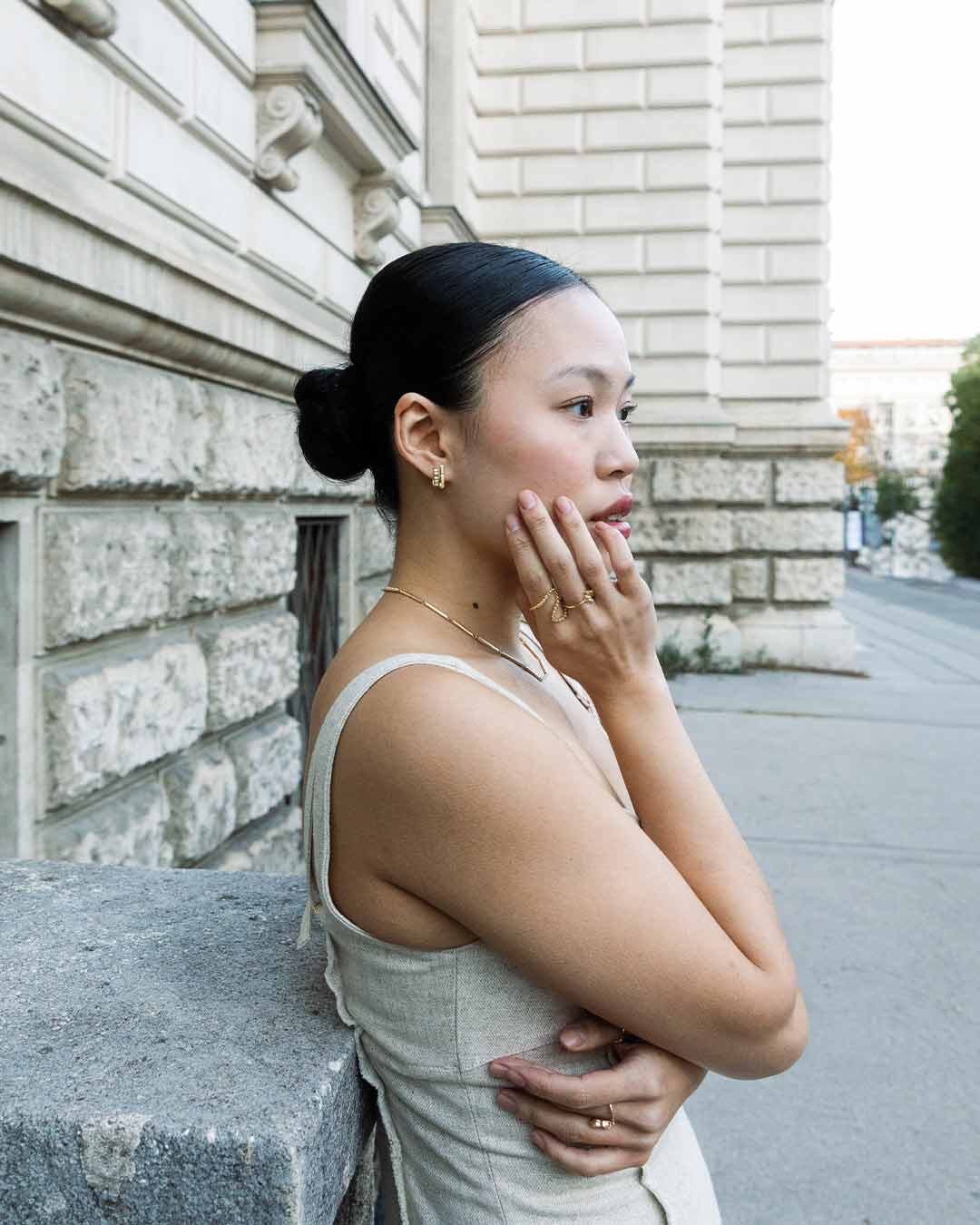 Asiatisches Model lehnt sich an eine neoklassizistische Fassade und wird von der Seite fotografiert wie sie achtsam nachhaltigen Schmuck von Maren Jewellery aus recyceltem Gold präsentiert